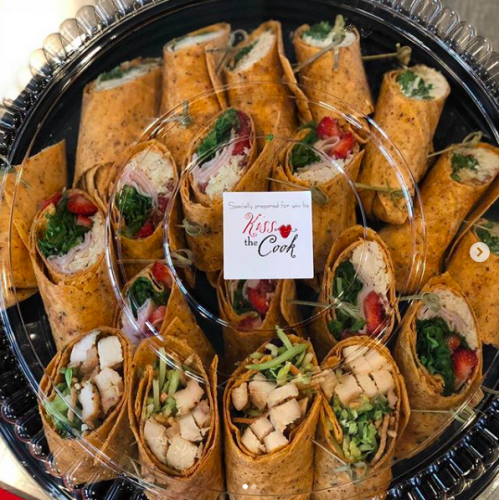 Tray of assorted rolled wraps with vegetables and chicken, garnished with greens and berries, in clear plastic packaging