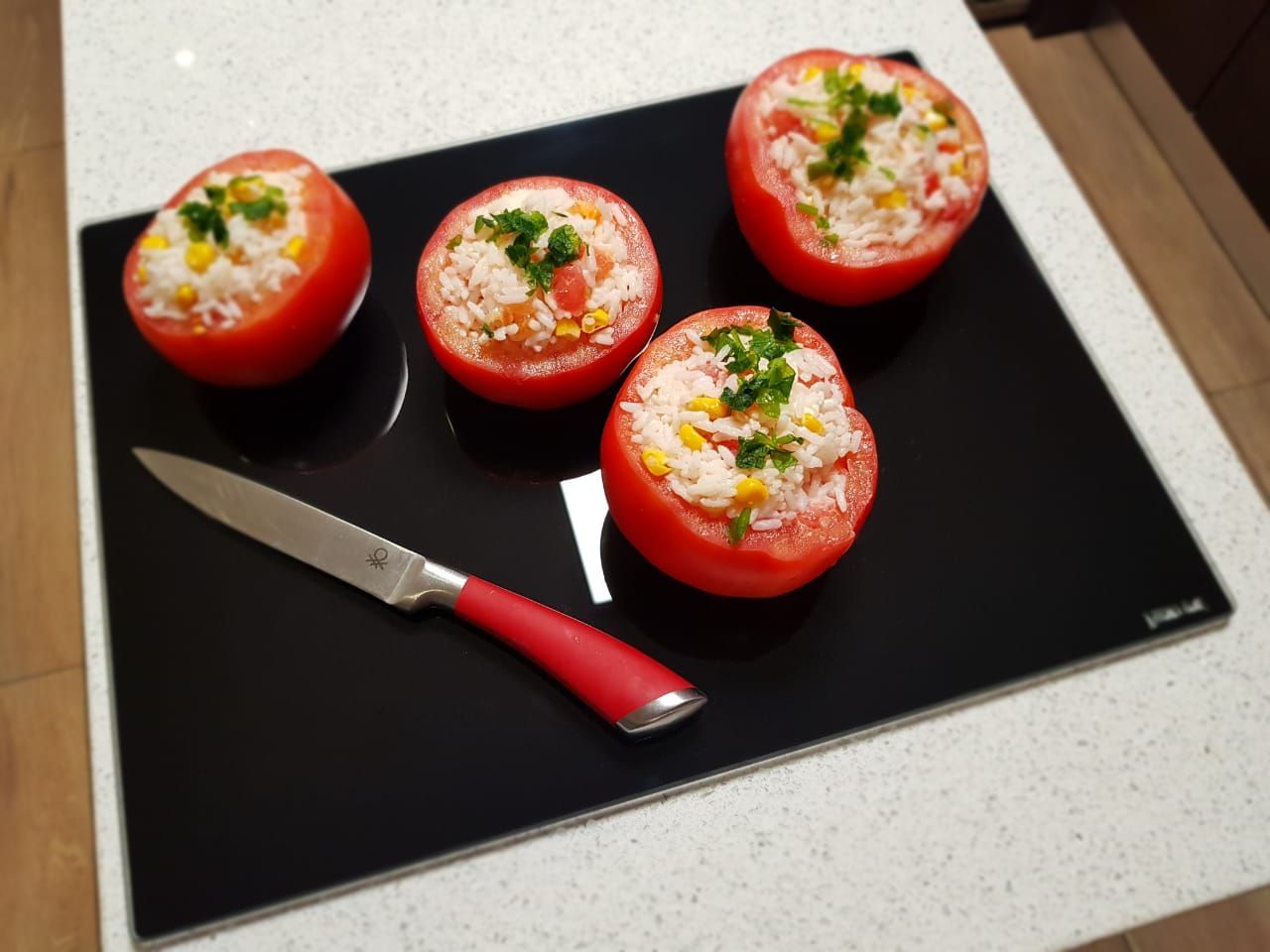 Cuatro tomates cortados por la mitad rellenos sobre una tabla de cortar negra, con cuchillo presente.
