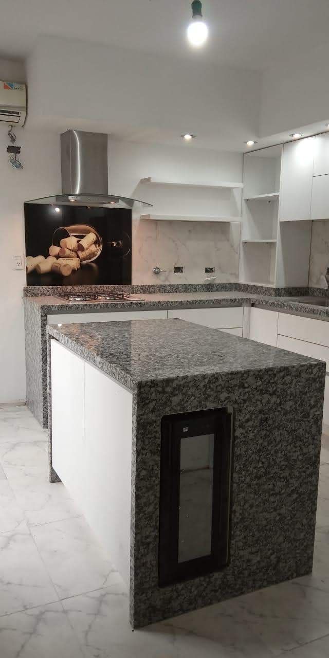 Cocina moderna con isla de granito y refrigerador empotrado. Muebles blancos y suelo de mármol.