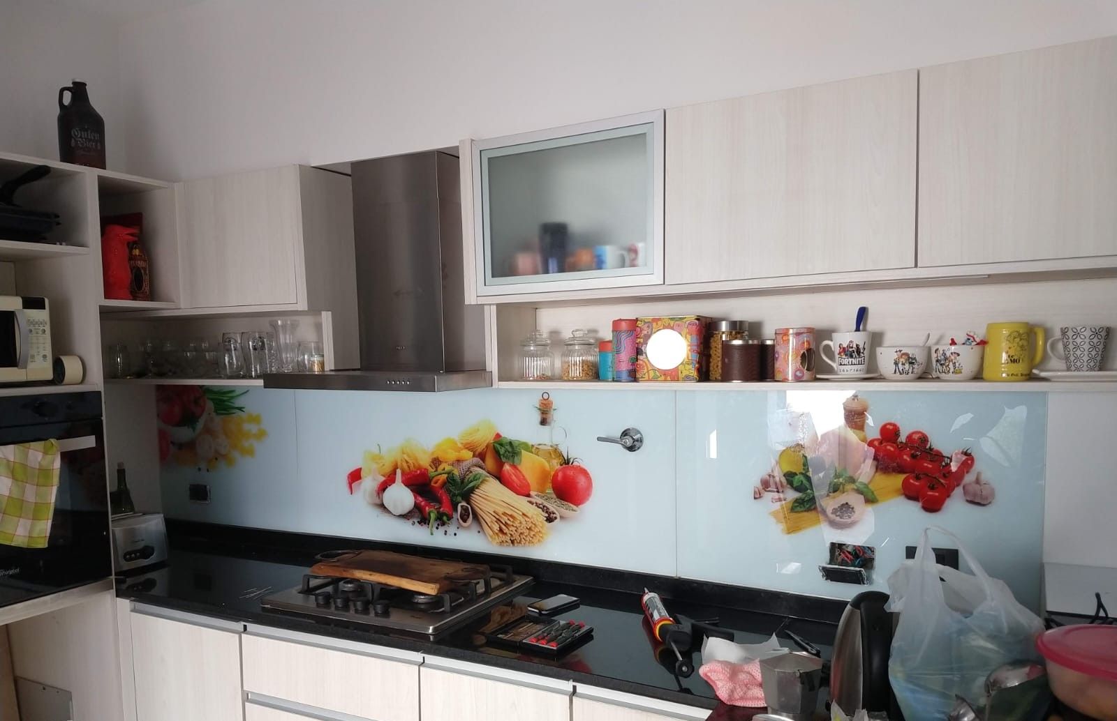 Cocina con gabinetes claros, encimera negra y protector contra salpicaduras de vidrio con imágenes de comida colorida.