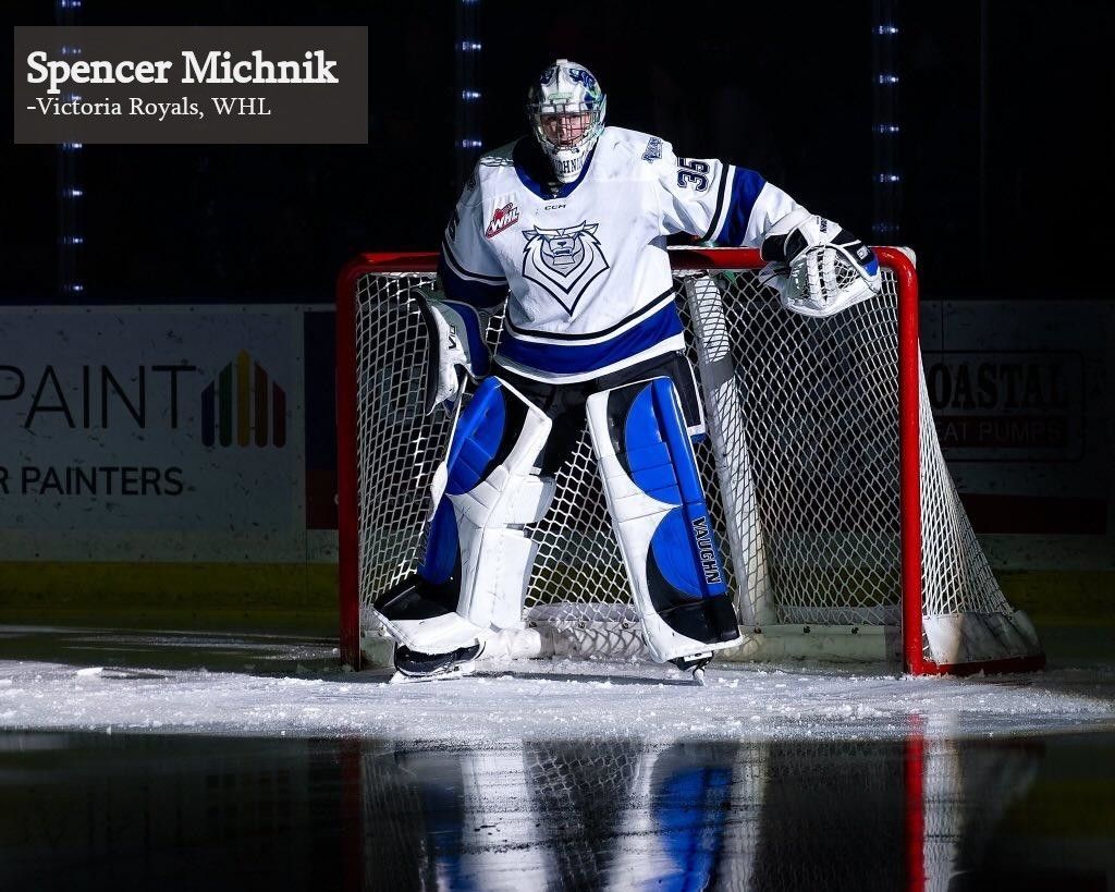 Victoria Royals hockey goalie Spencer Michnik stands in the net wearing white and blue gear on an indoor ice rink.