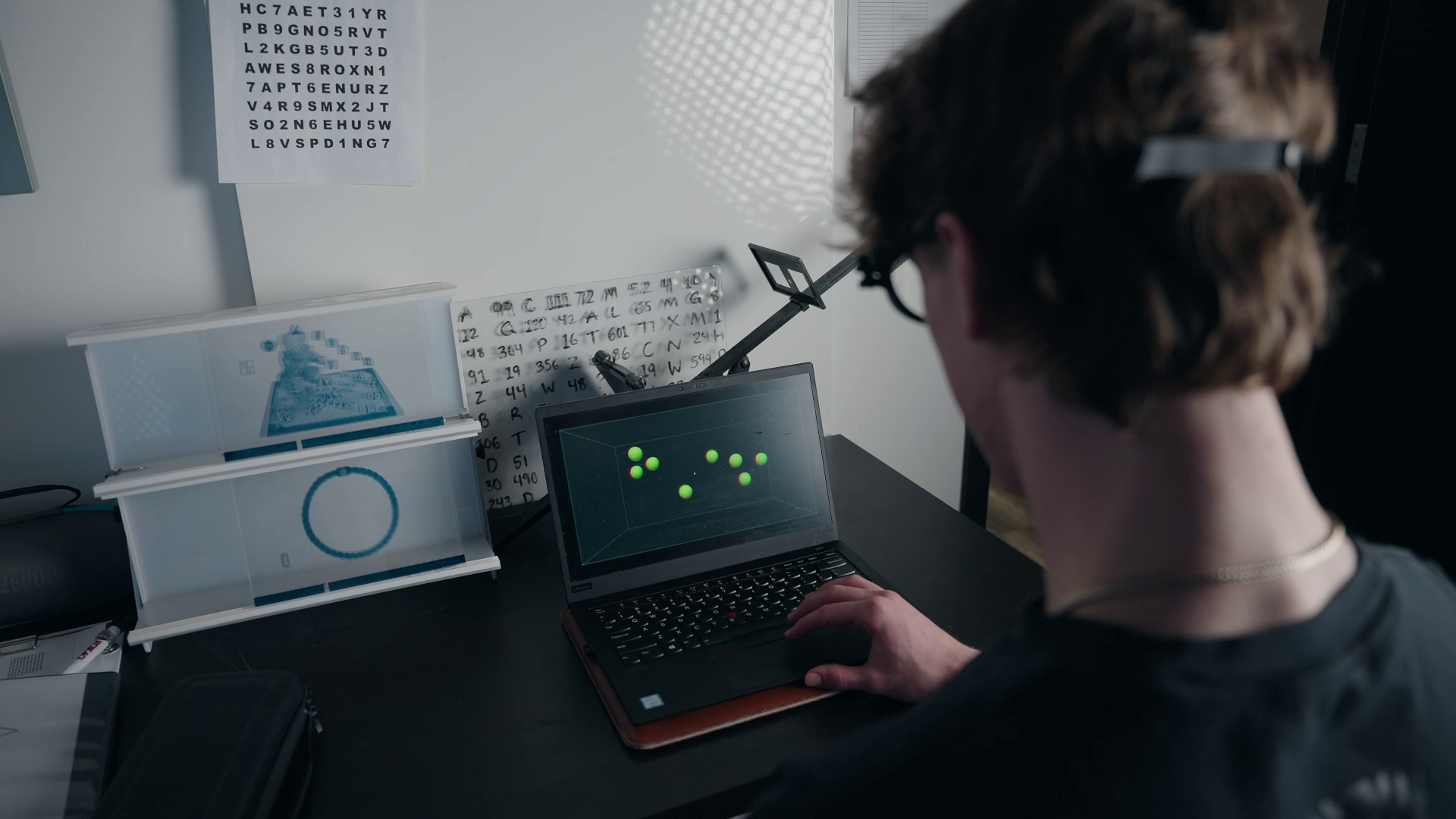 A person works at a desk on a laptop displaying green dots, with diagrams and a phone mount visible in the background.