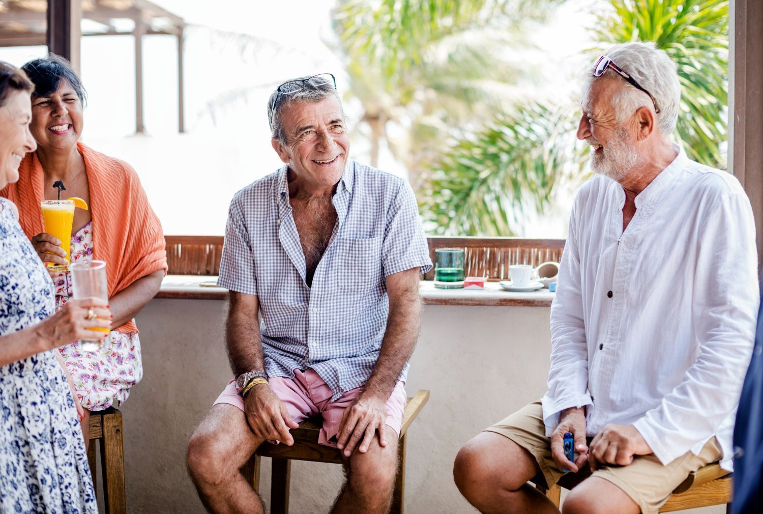 Group of people socializing on a balcony, two men smiling, one drinking, in a sunny setting.