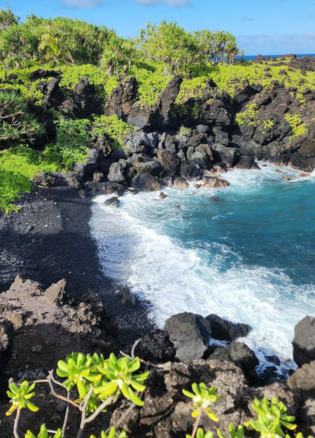 Black sand beach with foamy waves, dark rocks, green foliage, and clear blue water.