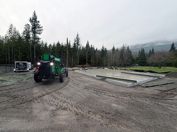 A green construction vehicle parked beside a concrete slab foundation on a dirt lot.