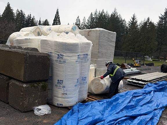 A worker in a high-visibility vest lifts a roll of insulation beside pallets of construction materials.