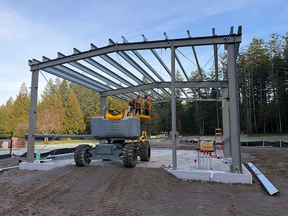 A construction crew works from a boom lift to assemble a steel frame building structure on a concrete foundation.