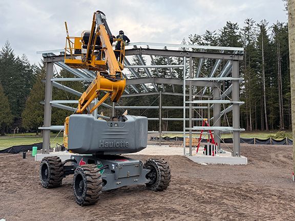 A yellow boom lift raises a worker to install the roof framing of a steel building.