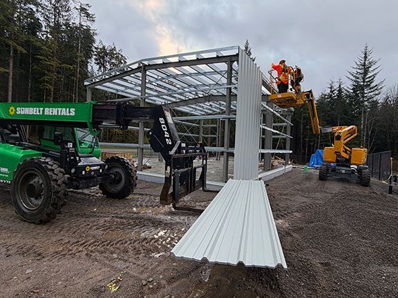 Construction workers and a boom begin to add metal wall panels to primary framing.