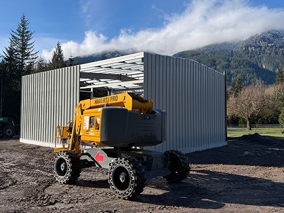 A yellow construction lift parked in front of a metal-walled building under construction against a mountain backdrop.