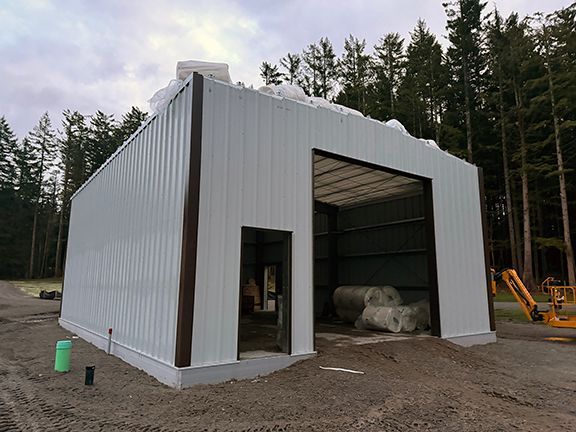 A white metal building under construction with a large open bay and a side door, situated in a wooded area.