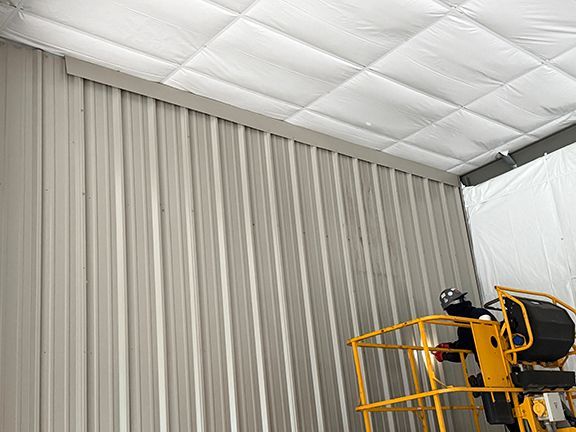 A worker in a yellow boom lift installs insulation panels on the corrugated metal wall and ceiling of an industrial space.