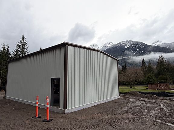 A light-colored metal storage building with a doorway, standing against a mountain and forest backdrop of B.C.