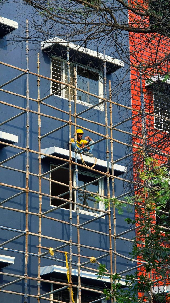 Worker in a yellow helmet on scaffolding beside a blue building with red brick trim