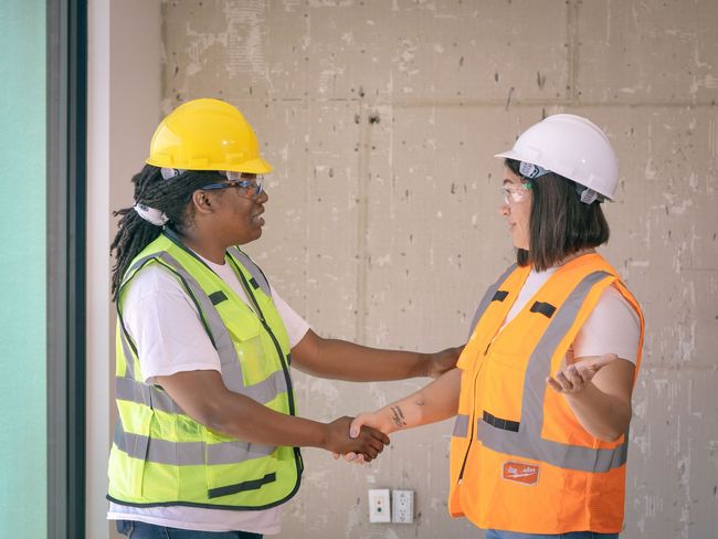 Two construction workers in safety vests and hard hats shake hands at a building site.