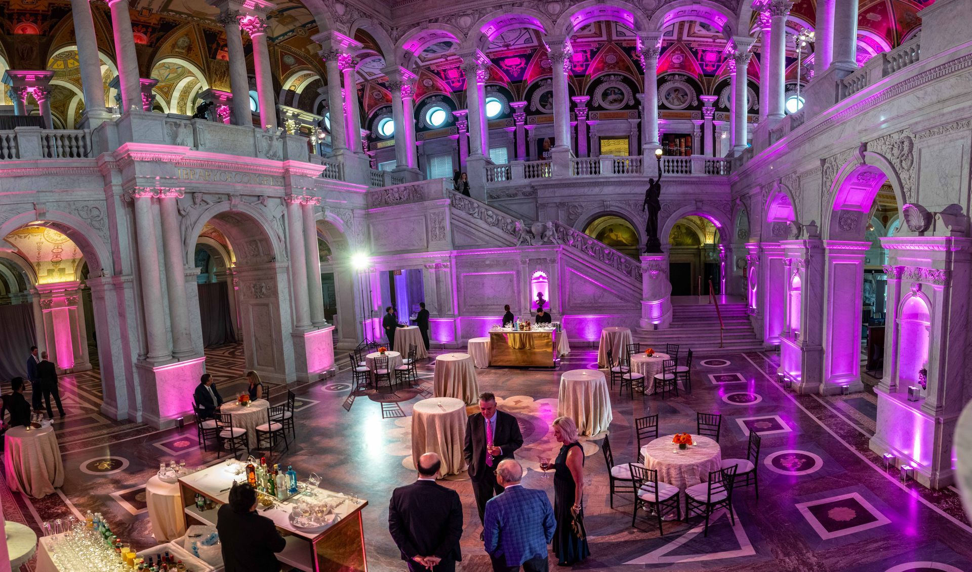 A group of people are standing in a large room with tables and chairs.