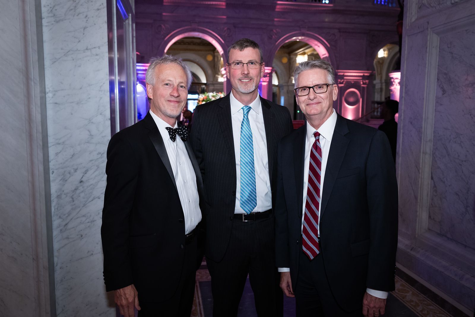 Three men in suits and ties are posing for a picture in a hallway.