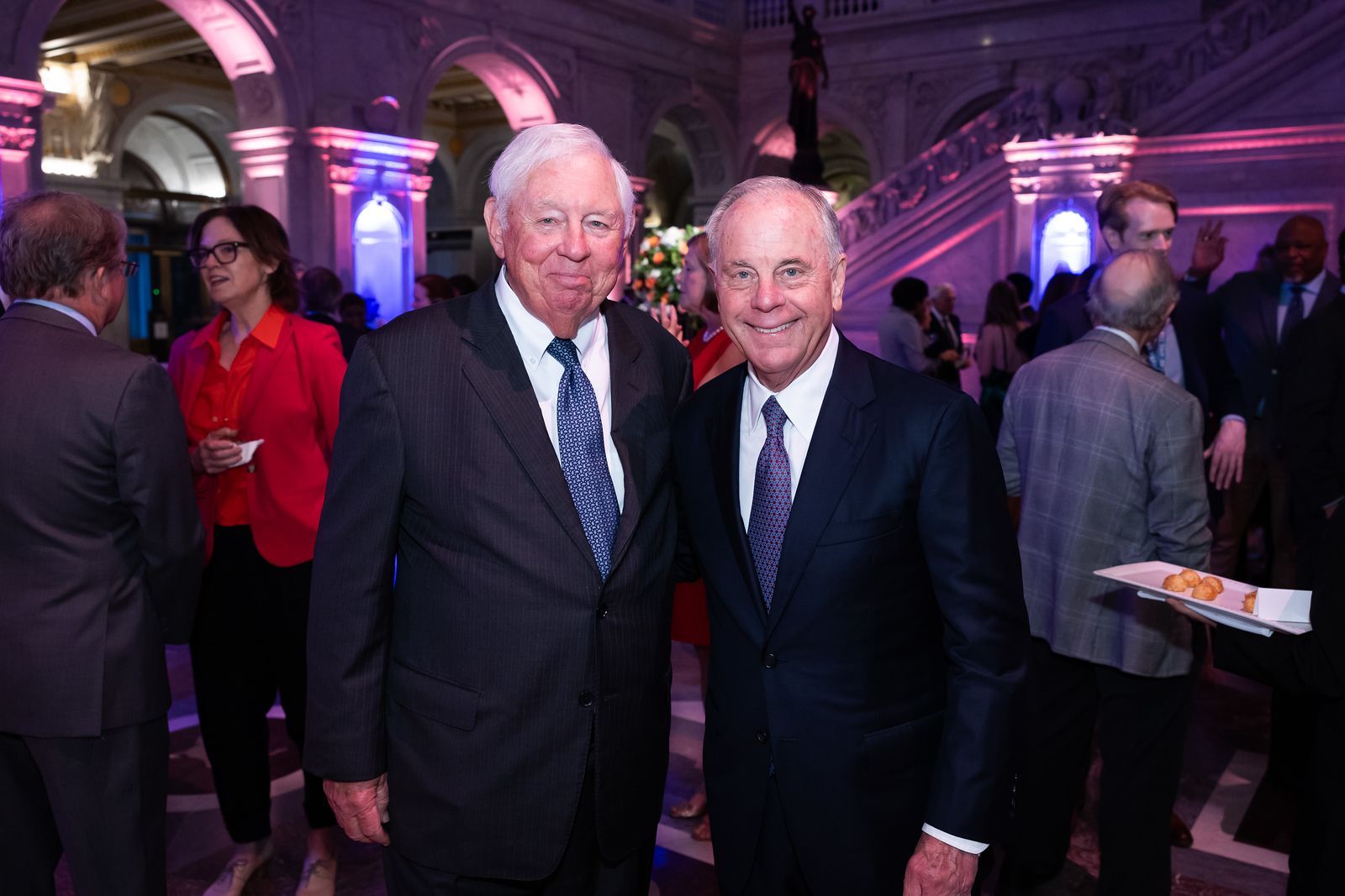 Two men in suits and ties are posing for a picture in a crowded room.