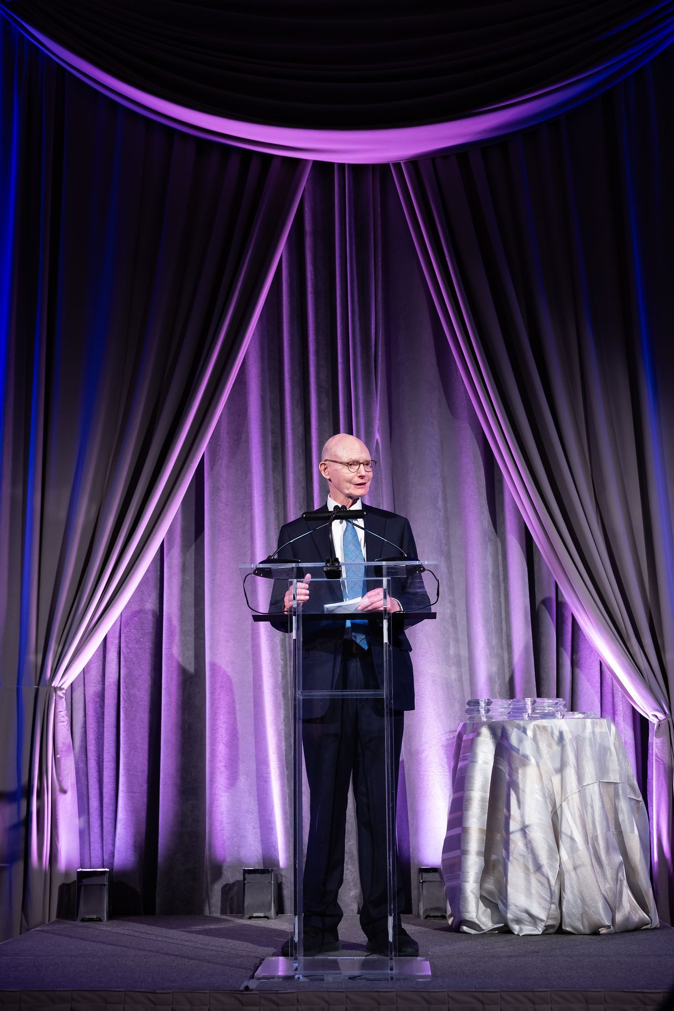 A man is standing at a podium giving a speech in front of a purple curtain.