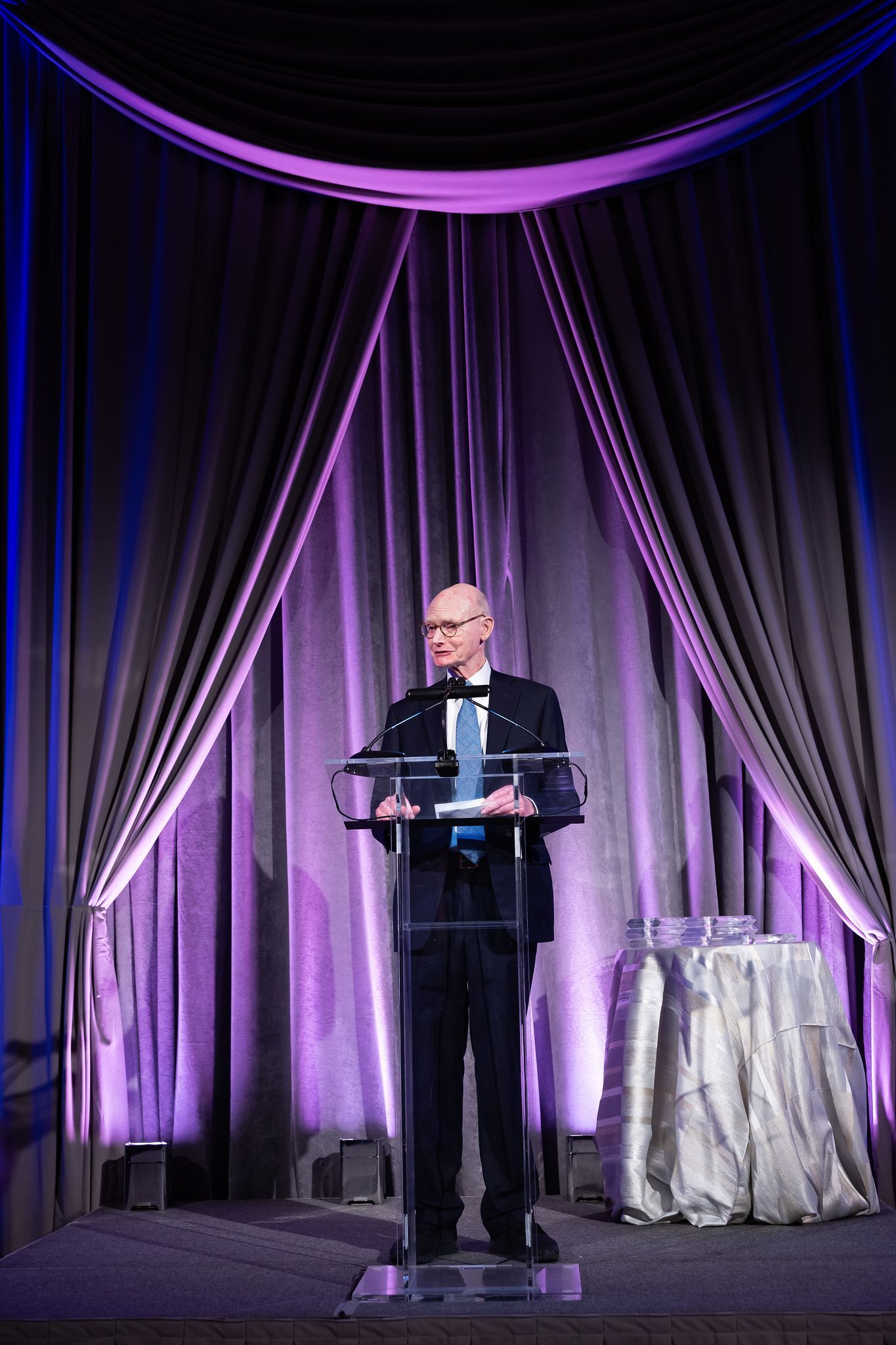 A man is standing at a podium giving a speech in front of a purple curtain.