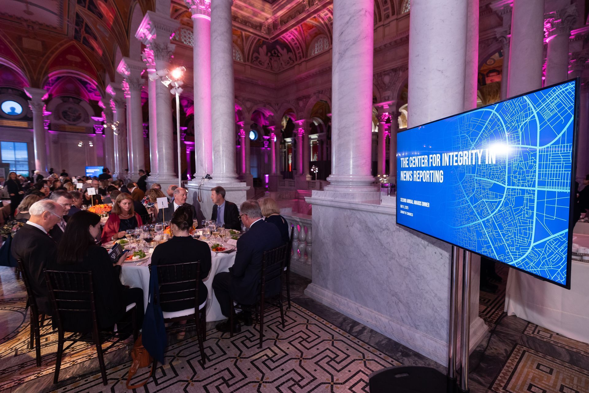 A group of people are sitting at tables in front of a large screen.