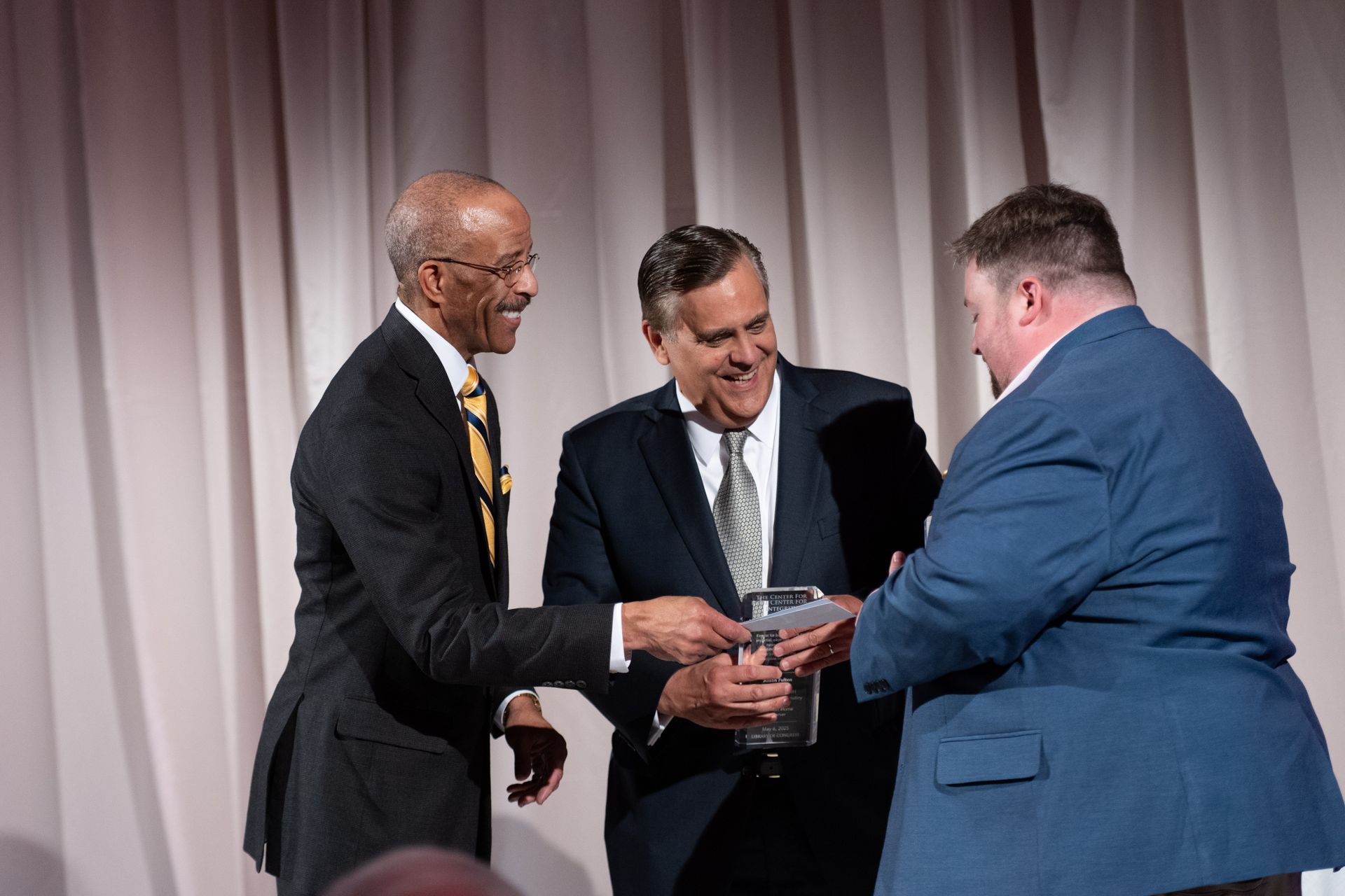 Three men in suits and ties are shaking hands on a stage.
