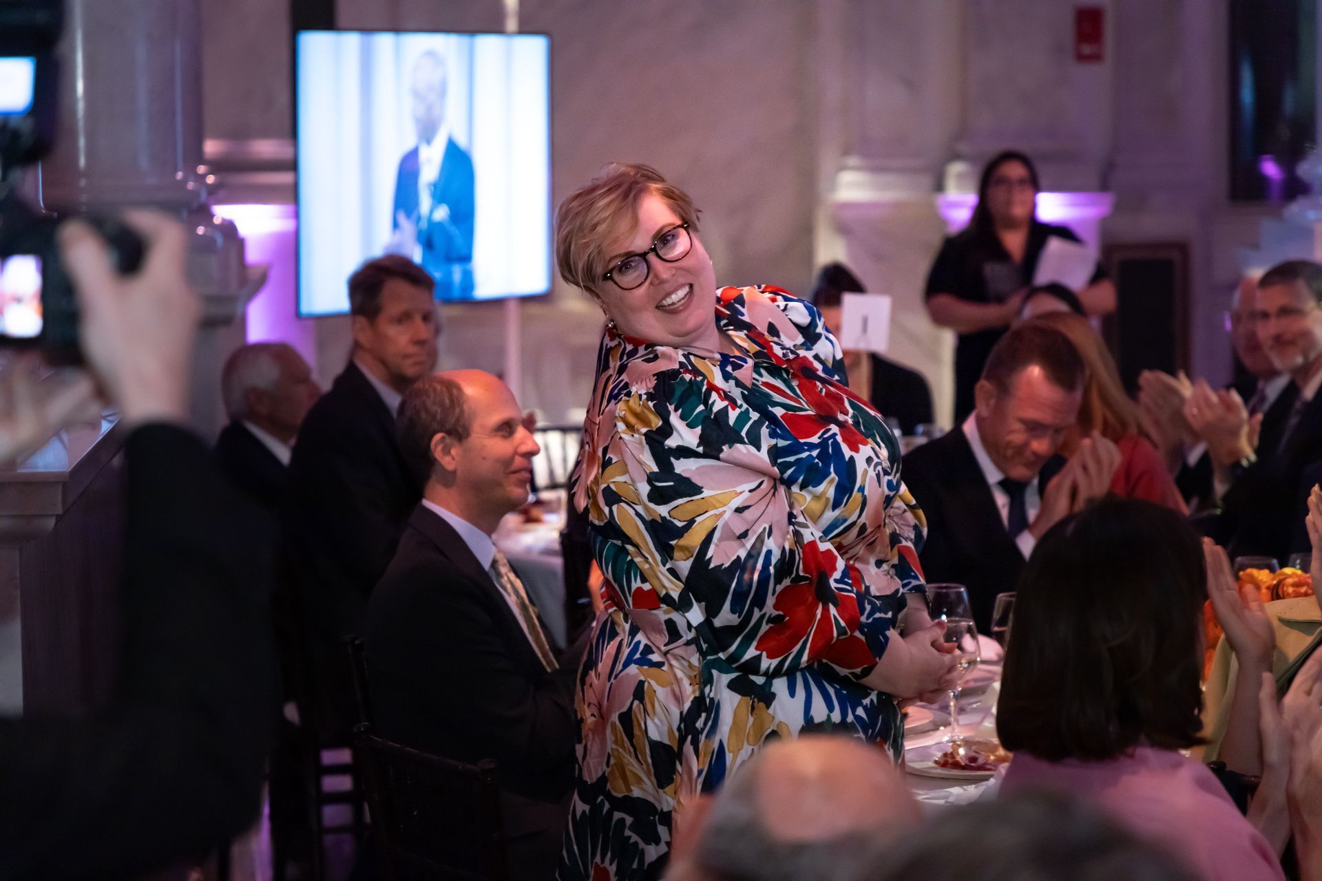 A woman in a floral dress is standing in front of a crowd of people at a party.