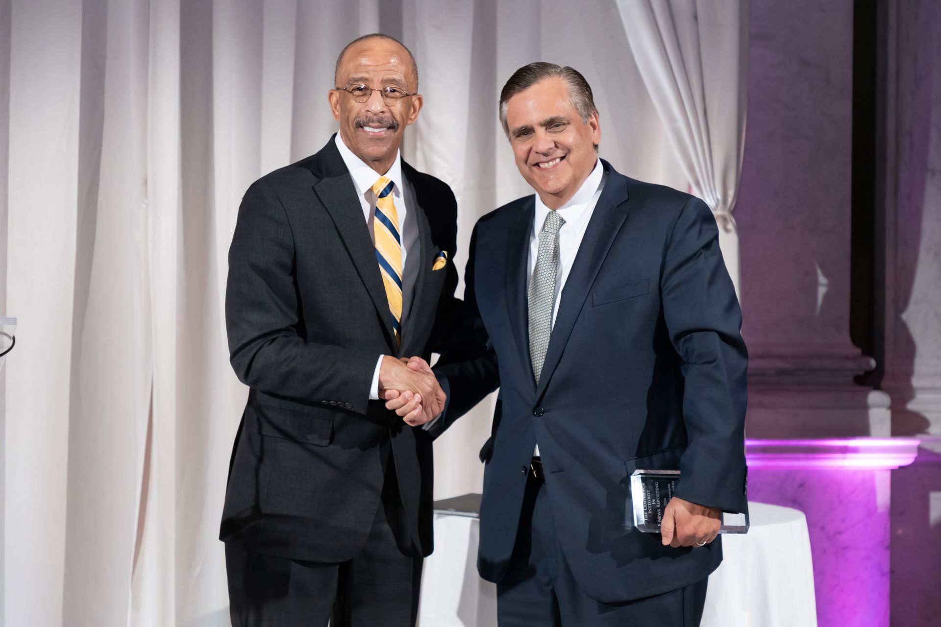 Two men in suits are shaking hands in front of a white curtain.