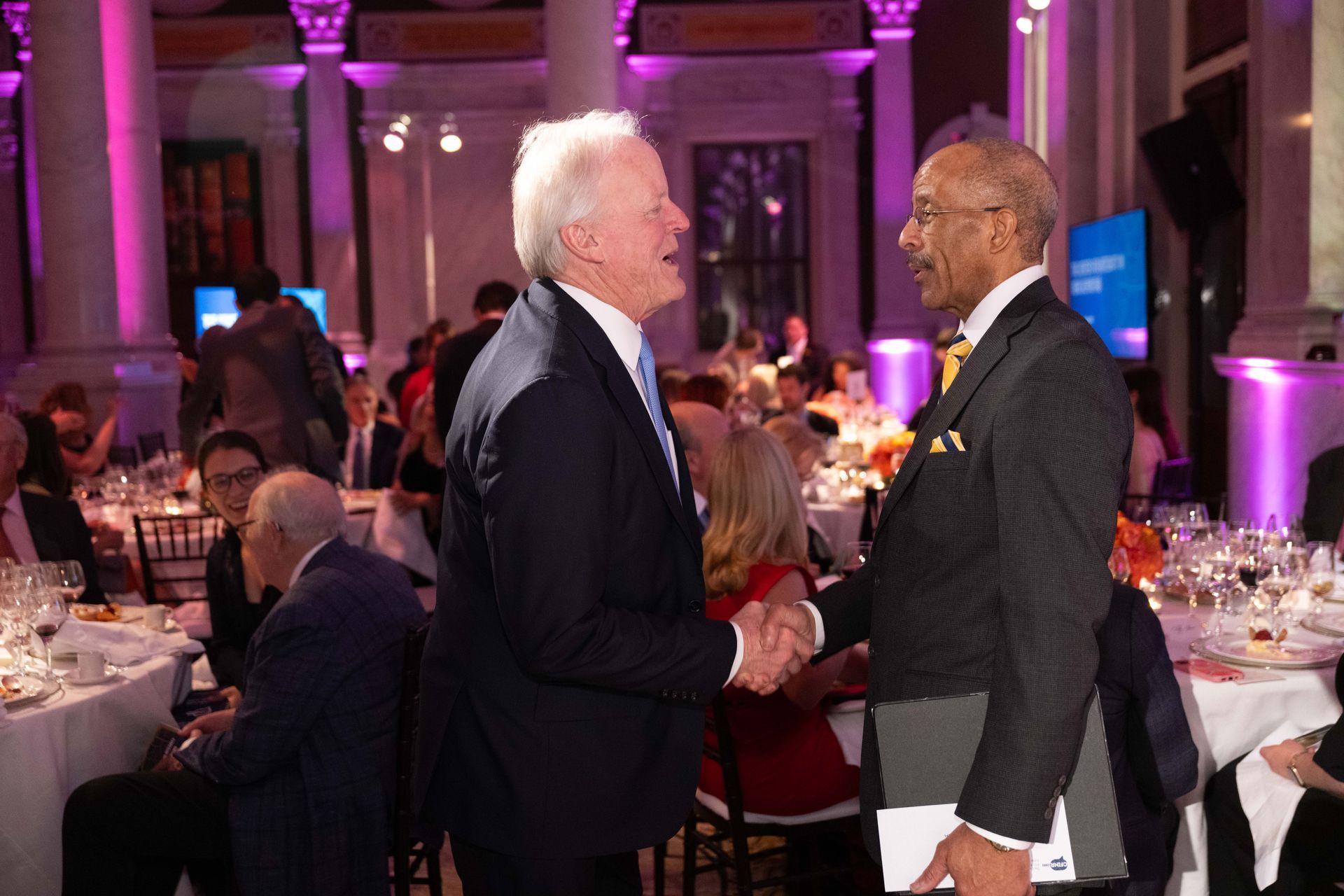 Two men are shaking hands in a room with purple lights.