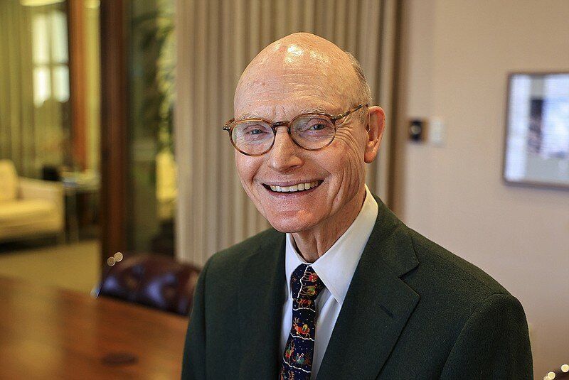 A man in a suit and glasses is smiling while talking into a microphone.