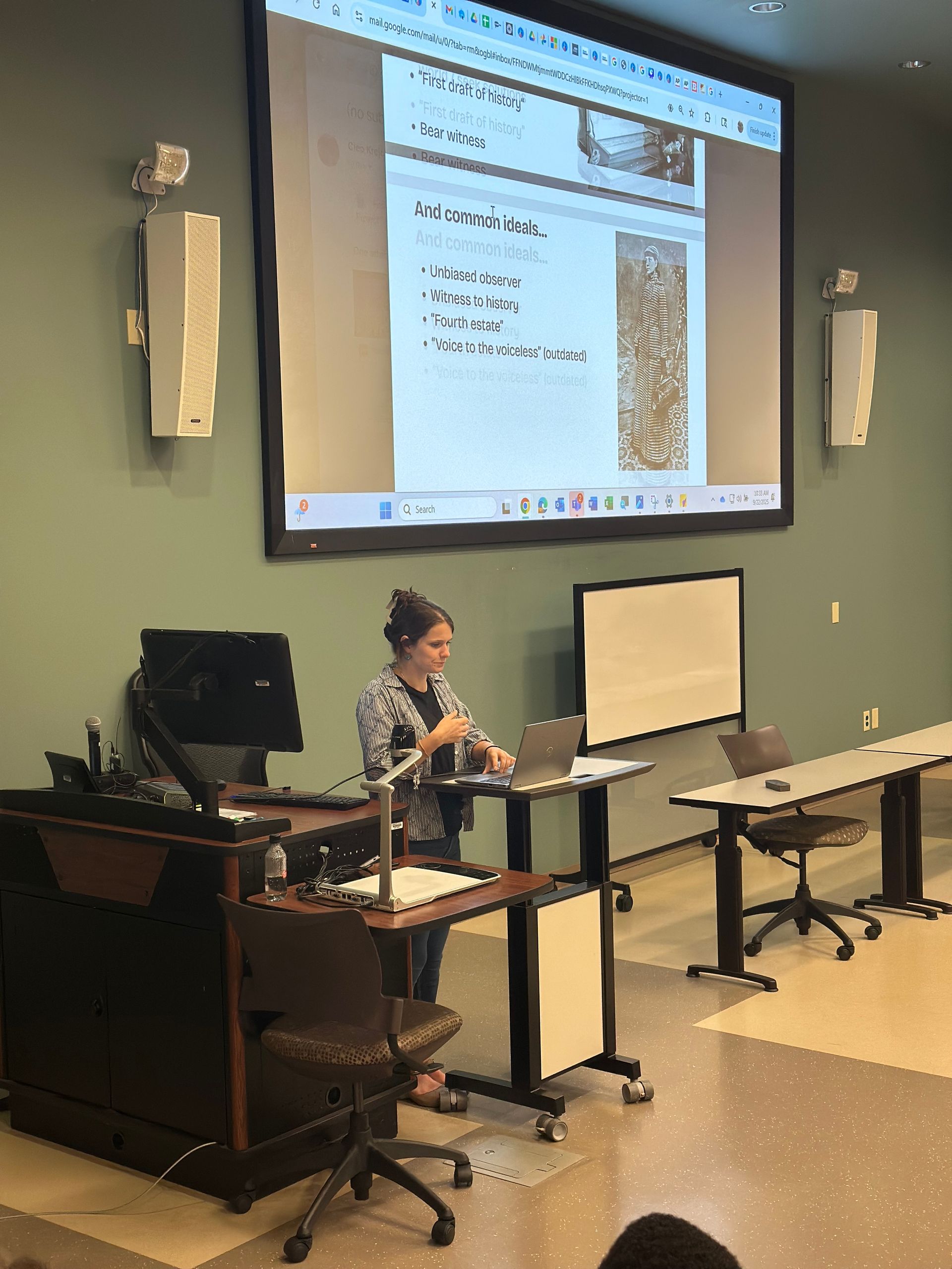 Woman giving presentation in a classroom, standing at a lectern. Slide on projector, whiteboard, tables, and chairs visible.