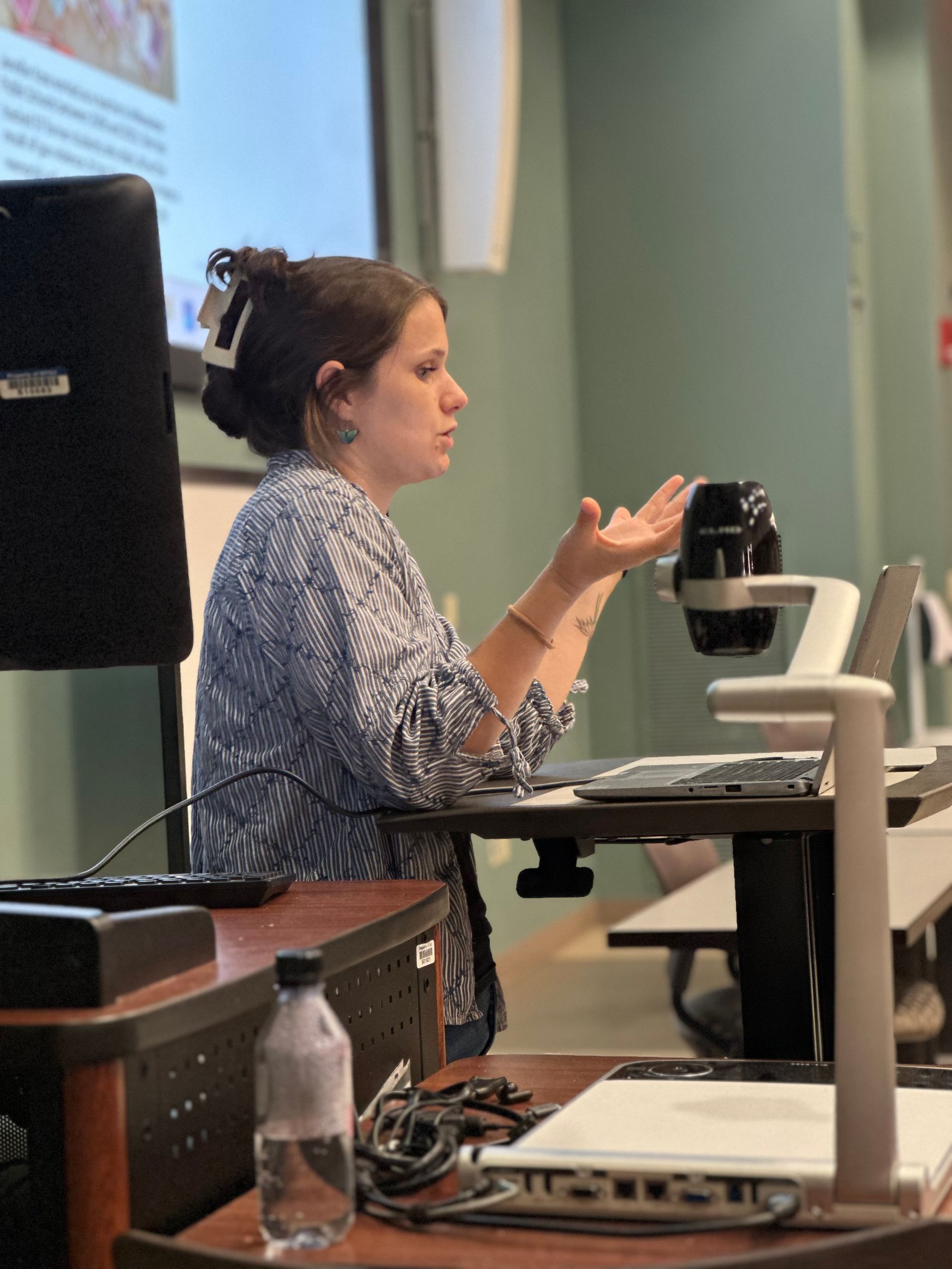 Woman gestures while teaching at a desk in a classroom. She has dark hair and wears a blue patterned top.