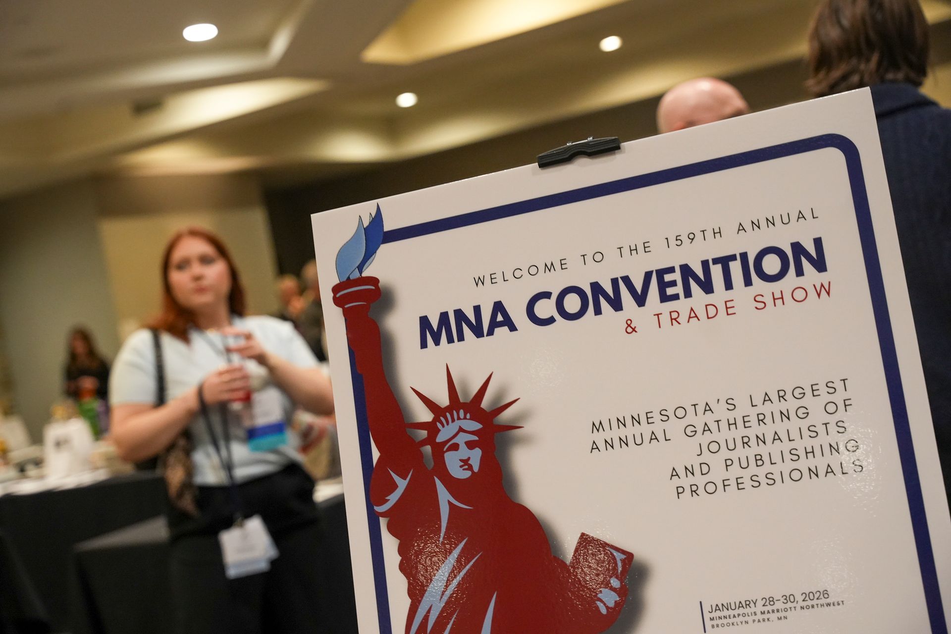 Woman gestures while teaching at a desk in a classroom. She has dark hair and wears a blue patterned top.