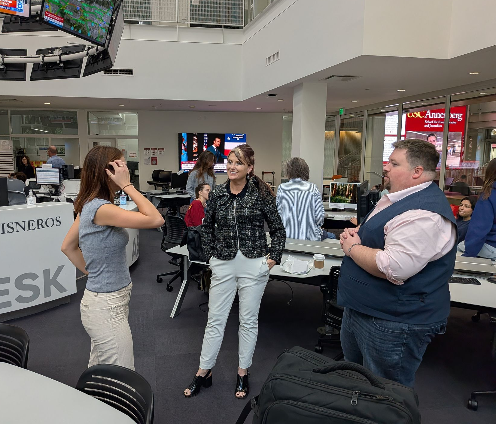 Three people converse at a modern desk with monitors; a newsroom setting. Two women and a man.