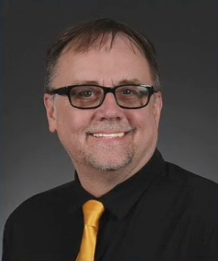 A man wearing glasses and a yellow tie is smiling for the camera.