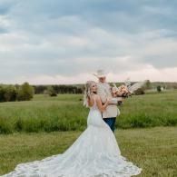 Bride in a white gown standing in a grassy field with a bouquet beside a person holding flowers under a cloudy sky