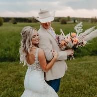 Bride hugging partner in a white cowboy hat, holding a bouquet in a grassy field at sunset