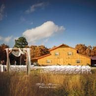 Outdoor wedding setup with white chairs and draped arch in a field beside a yellow barn under a blue sky