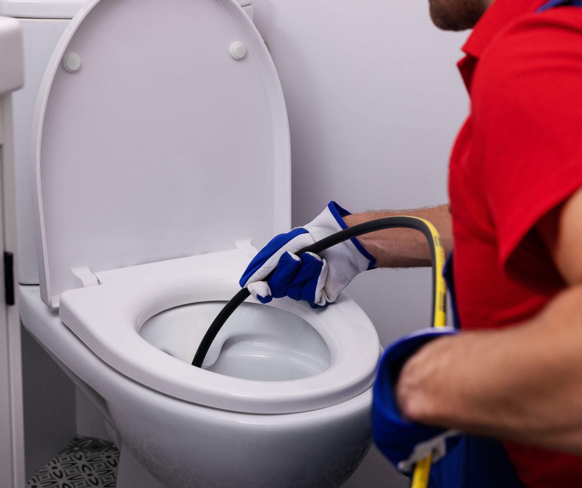 Person unclogs a toilet with a snake tool, wearing gloves, in a bathroom.