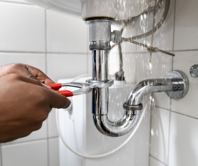 Person using pliers on a leaking chrome sink pipe in a white bathroom.