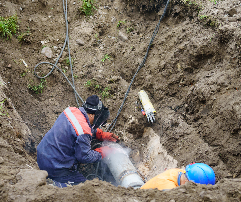 Workers welding a pipe in a trench. One wears a welding helmet, the other a hard hat.