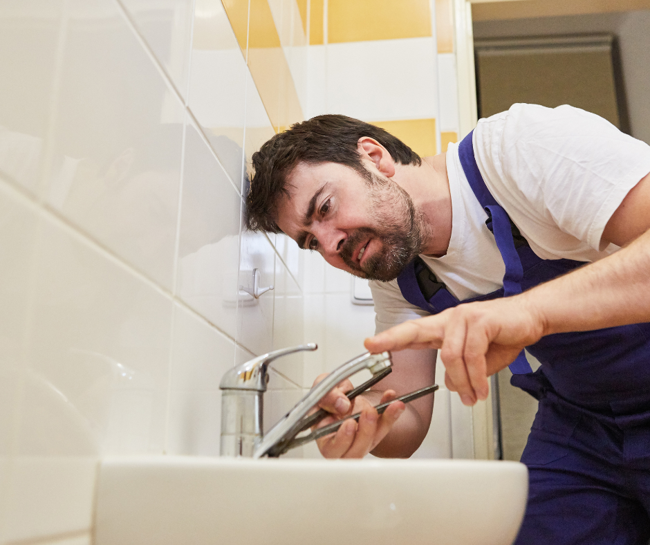 Plumber in blue overalls working on a sink faucet in a bathroom.