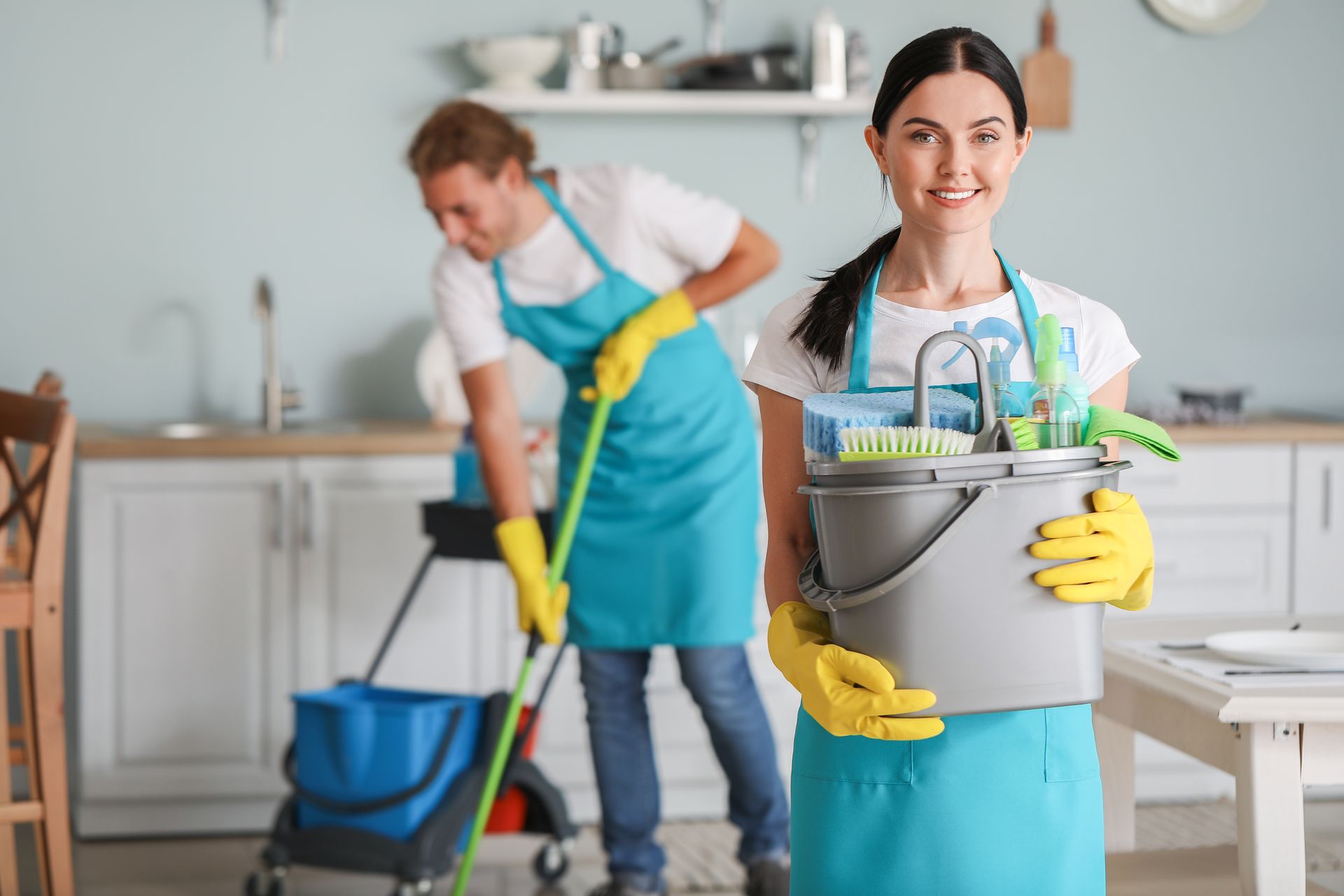 A female janitor with cleaning supplies in a kitchen. A male janitor mops in the background.