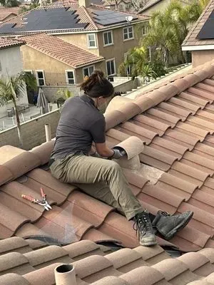 A worker in a gray shirt and tan pants kneels on a brown tiled roof, installing a vent cover with a hand tool.