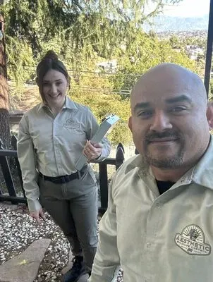 Two professionals in tan uniforms pose outdoors on a sunny day; one holds a clipboard, and both smile at the camera.