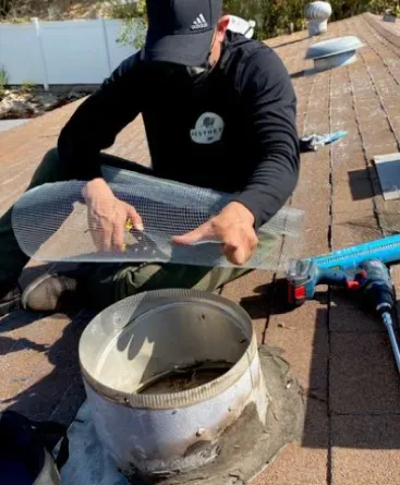 A person in a black cap and shirt kneeling on a roof, cutting wire mesh to cover a circular roof vent.