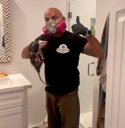 A bald person in a respirator and black gloves holds an opossum in a bathroom while giving a thumbs-up.