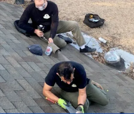 Two people in black shirts work on a shingled roof, kneeling and using tools to repair a flashing section.