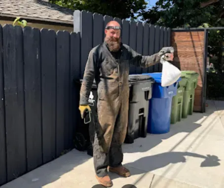 A smiling person in mud-stained work coveralls holding a white bag near a row of trash bins against a dark fence.