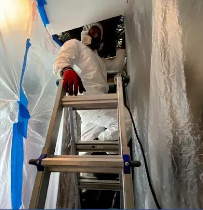 A person in a full white protective suit and respirator mask stands on a ladder inside a plastic-enclosed work area.